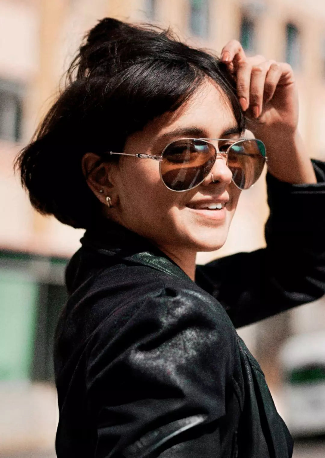 Mulher com cabelo curto e franja usando óculos de sol estilo aviador com lentes marrons. Ela veste uma jaqueta preta e sorri, destacando um look casual e urbano em ambiente externo.