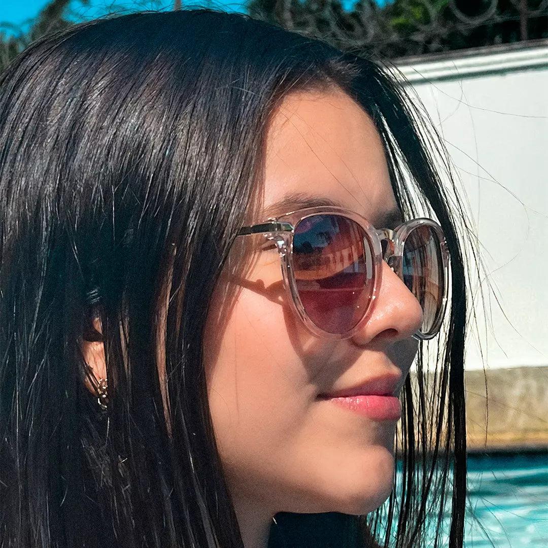 Mulher usando óculos de sol com armação transparente e lentes degradê rosadas, cabelo preto solto e fundo com piscina em dia ensolarado. Estilo delicado e moderno para ocasiões ao ar livre.